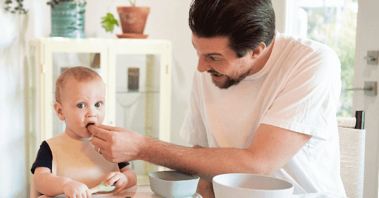 A smiling father enthusiastically feeds his infant son at a dining table.