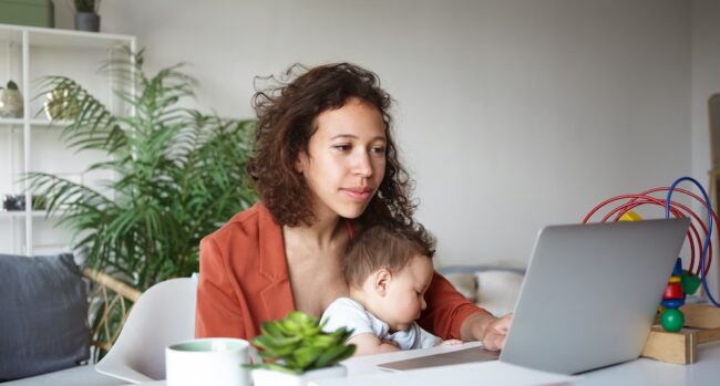 Motherhood, career and employment concept. Casually dressed young mother nursing baby and looking for job online, browsing websites, sitting at desk in front of open laptop