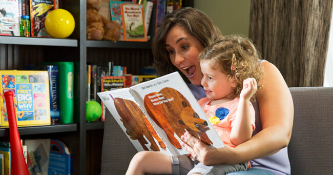 Mom with a preschool-age girl in her lap, reading a book.